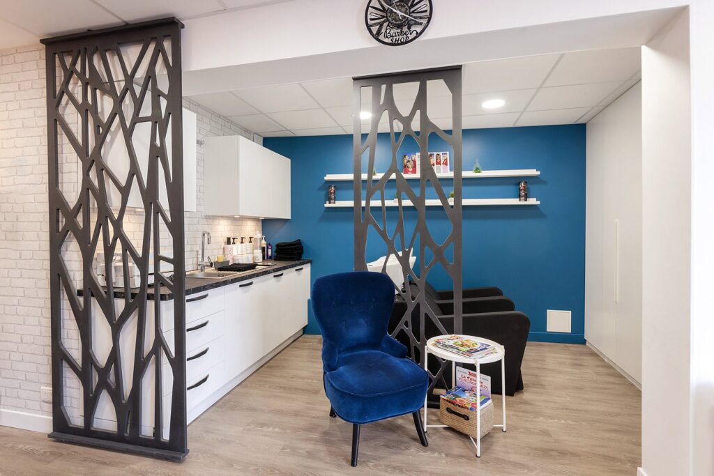Intérieur de salon moderne avec deux chaises noires, un fauteuil bleu, une petite table avec des magazines, des cloisons décoratives et des armoires blanches contre un mur d'accent bleu.