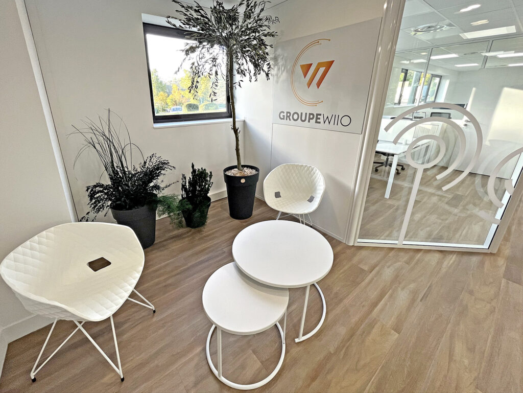 Petit salon de bureau avec deux chaises blanches, des tables rondes gigognes, des plantes en pot et un mur avec le logo "GROUPE WIIO" ; cloison en verre à l'arrière-plan.