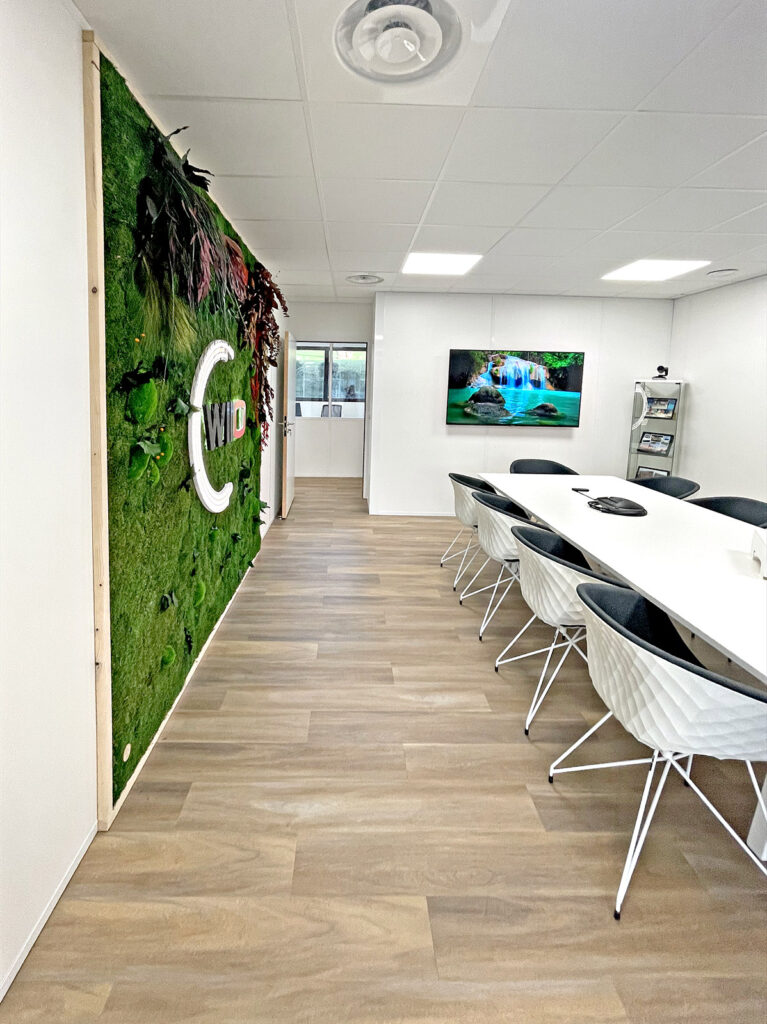Salle de conférence moderne avec une longue table blanche, des chaises noires et blanches, un mur de mousse verte avec les lettres "CWD" et un écran mural affichant une image de chute d'eau.
