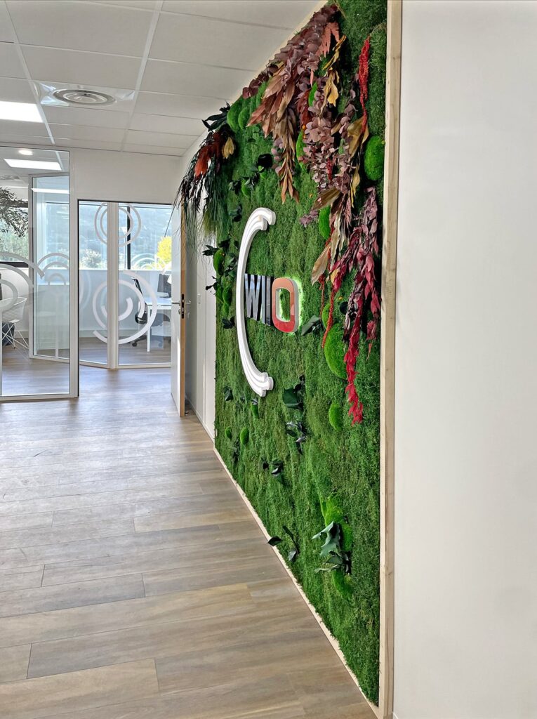 Un couloir avec un mur de mousse verte sur lequel figure le logo d'une entreprise, encadré par des bordures blanches et décoré de feuilles et de plantes séchées. Des portes de bureau en verre sont visibles à l'arrière-plan.