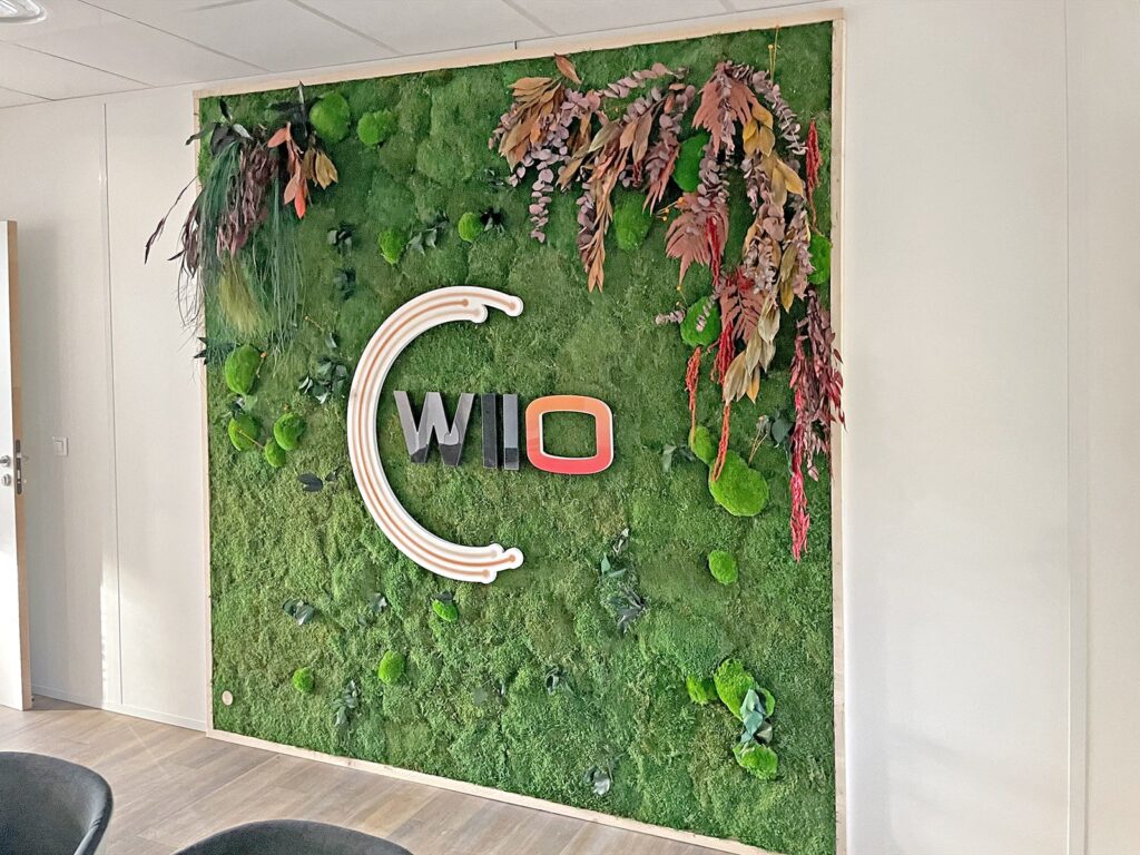 Un mur de mousse verte avec le texte "WILO" en lettres d'imprimerie et une forme ovale rose, décoré de plantes séchées suspendues et d'accents de bois clair.