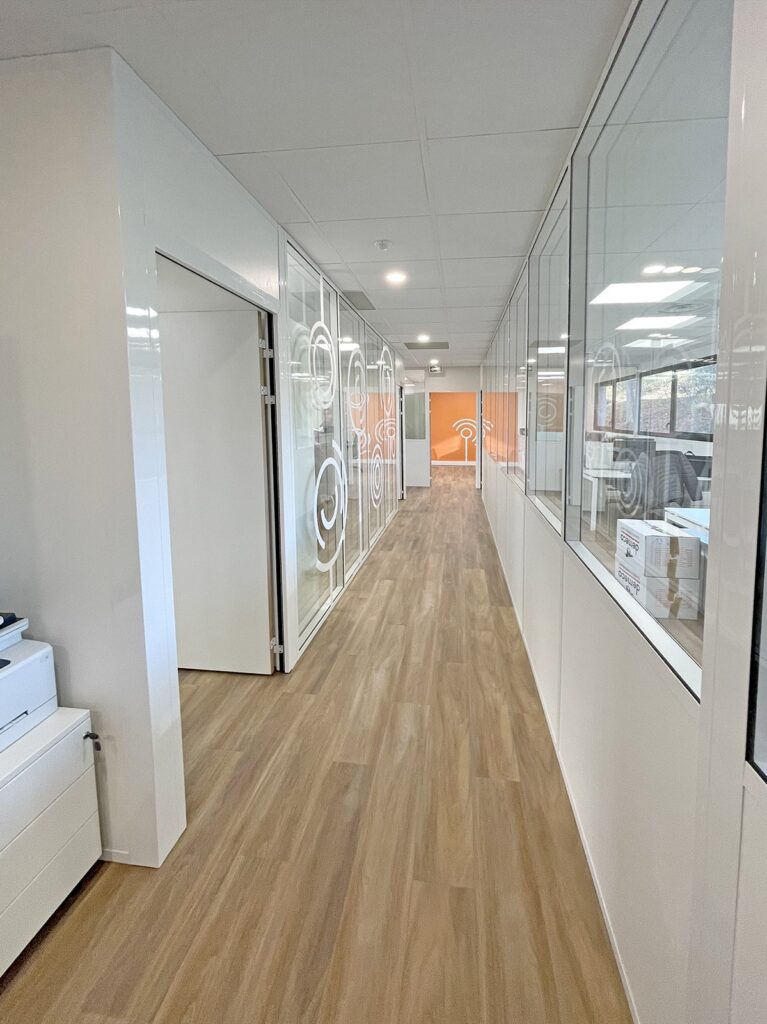 Un couloir de bureau moderne avec des pièces vitrées à droite et un mur blanc avec une porte à gauche.