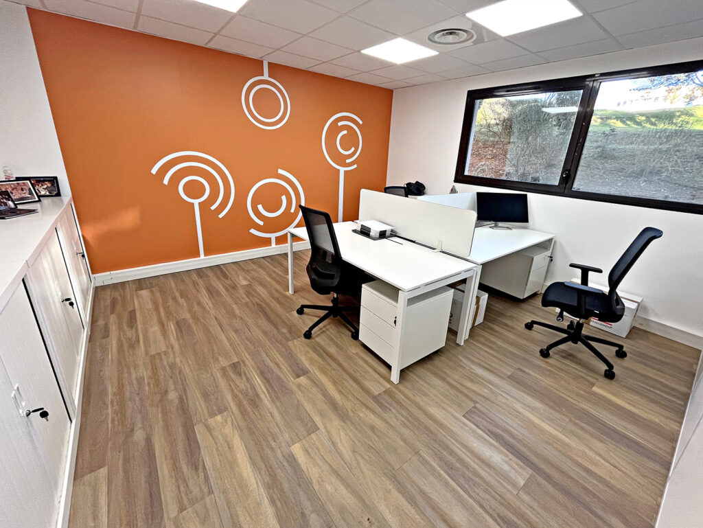 Bureau moderne avec deux bureaux, des chaises noires, des ordinateurs, un sol en bois, un mur d'accent orange avec des motifs abstraits blancs et une grande fenêtre apportant de la lumière naturelle.