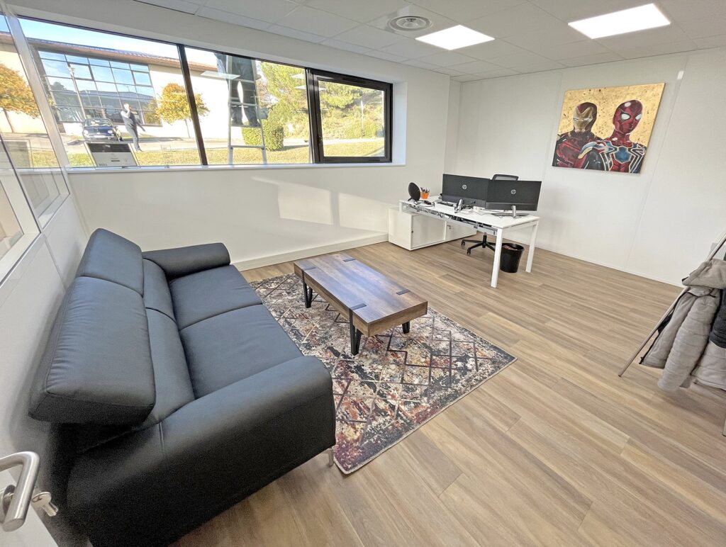 Bureau moderne avec un canapé noir, un tapis à motifs, une table basse en bois, deux bureaux avec écrans, une grande fenêtre et un tableau mural représentant deux personnages de bande dessinée.