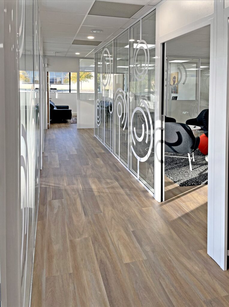 Un couloir de bureau moderne avec un sol en bois, des murs en verre avec des motifs circulaires et un coin salon avec des chaises noires visibles à travers une porte ouverte.
