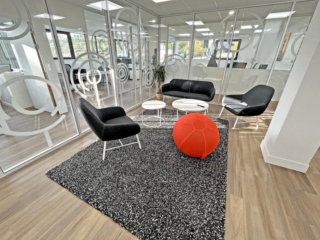Salon de bureau moderne avec des murs en verre, des chaises noires, un siège sphérique rouge, des tables rondes et un tapis noir moucheté sur un sol en bois clair.