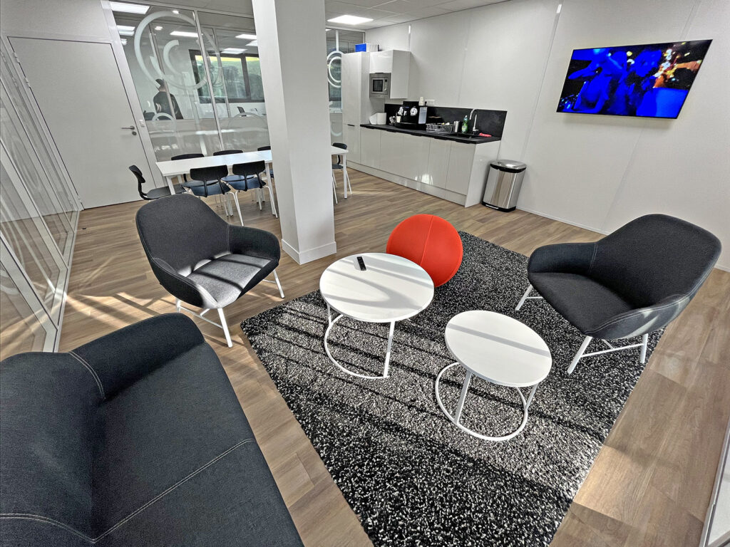 Salon de bureau moderne avec des chaises noires, un tabouret rouge, des tables rondes blanches, un tapis, une kitchenette, un téléviseur et une salle de réunion vitrée en arrière-plan.