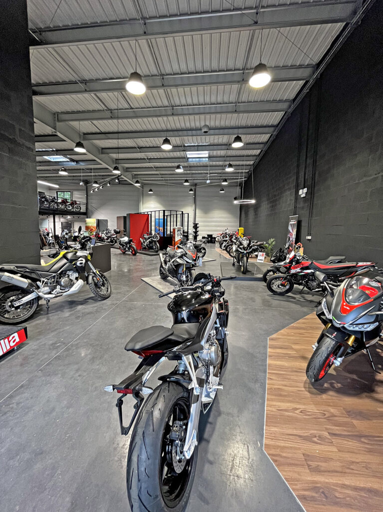 Une salle d'exposition de motos avec plusieurs motos exposées, avec un haut plafond, un sol en béton et des murs noirs sous un éclairage zénithal brillant.