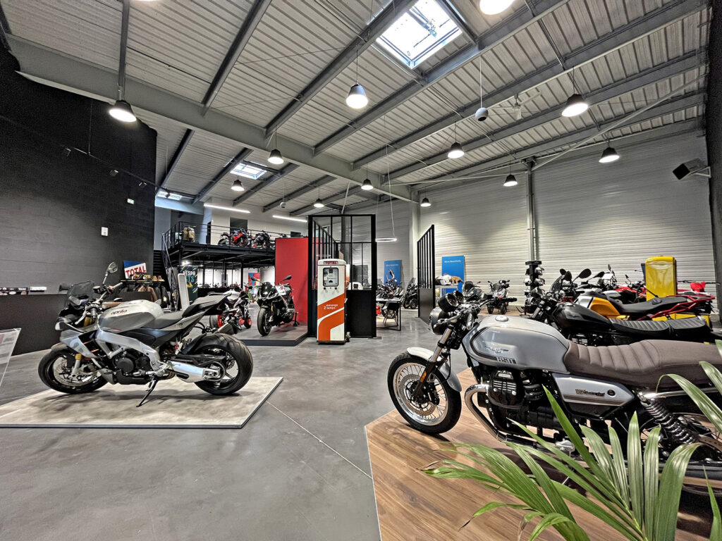 Intérieur d'une salle d'exposition de motos moderne avec plusieurs motos exposées, de hauts plafonds, un éclairage industriel et une mezzanine à l'arrière-plan.