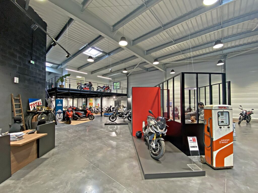 Salle d'exposition de motos avec plusieurs motos exposées, une pompe à essence d'époque et une mezzanine avec d'autres motos. Intérieur de style industriel avec de hauts plafonds et de grandes fenêtres.
