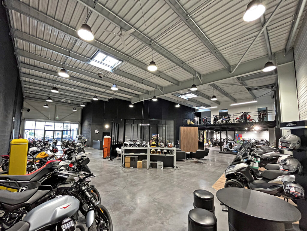 Salle d'exposition de motos moderne avec plusieurs motos exposées, des casques sur des tables et une mezzanine supérieure contenant d'autres motos.