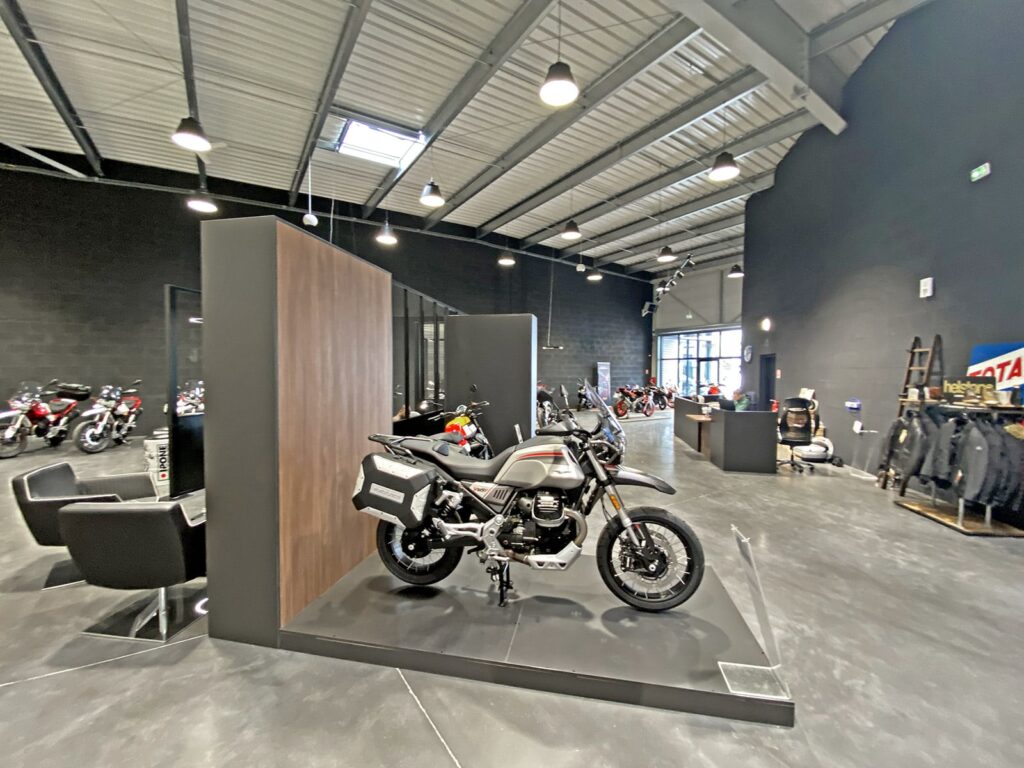 Une moto moderne est exposée sur une plate-forme à l'intérieur d'une salle d'exposition spacieuse, avec plusieurs autres motos et accessoires visibles à l'arrière-plan.