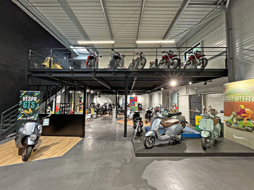 Une salle d'exposition présente divers scooters Vespa au rez-de-chaussée et à la mezzanine, avec des panneaux et des affiches Vespa colorés sur les murs.