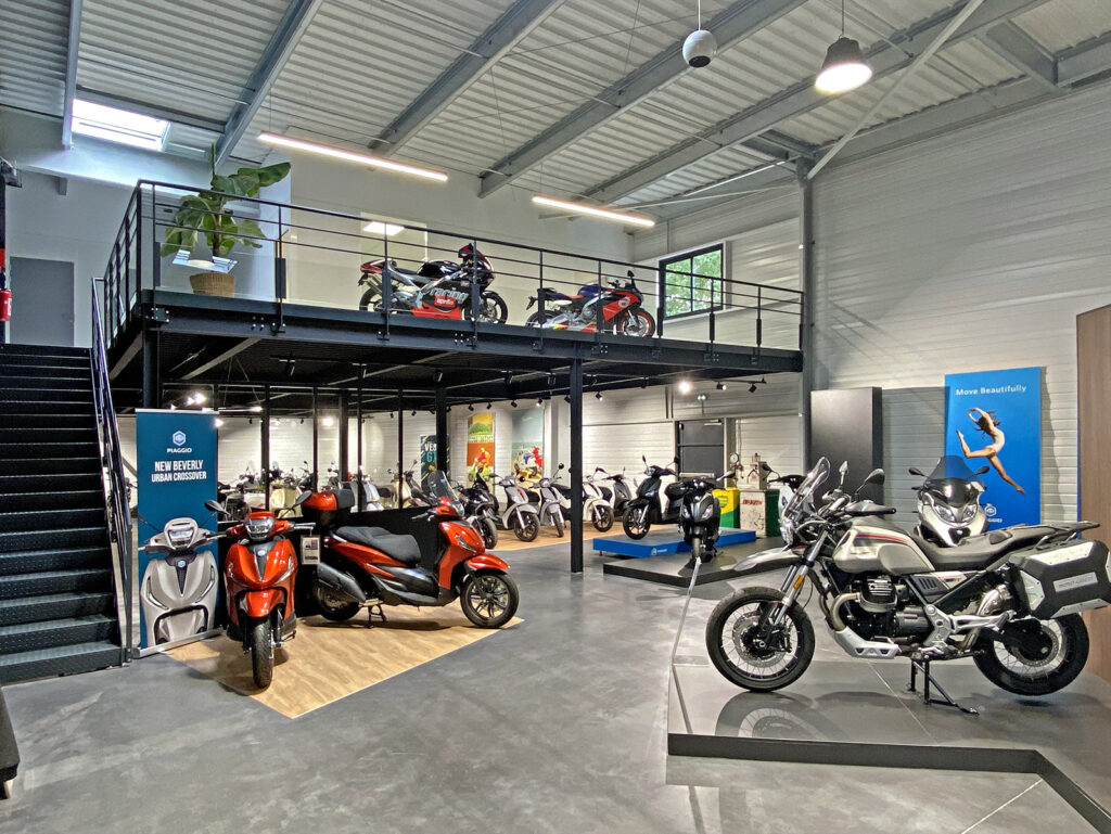 Une salle d'exposition de motos moderne avec différents modèles exposés au sol et sur la mezzanine, un éclairage vif et des affiches sur les murs.