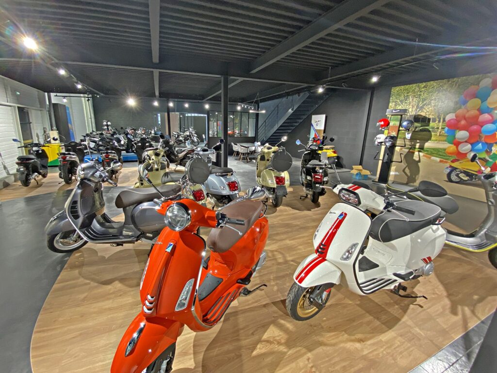 Une salle d'exposition présentant plusieurs scooters de différentes couleurs, dont certains sont garés sur une plate-forme en bois et d'autres sur un sol noir, sous des plafonniers lumineux.
