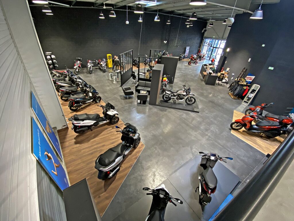 Intérieur d'une salle d'exposition avec plusieurs motos et scooters exposés, un éclairage vif et des accessoires qui tapissent les murs.