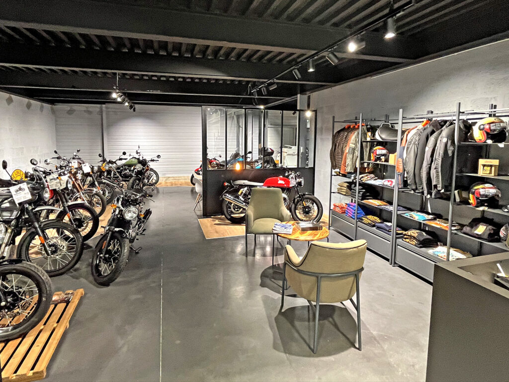 Une salle d'exposition de motos avec plusieurs motos exposées, deux chaises et une table au centre, et des racks de vestes et d'accessoires sur le mur de droite.