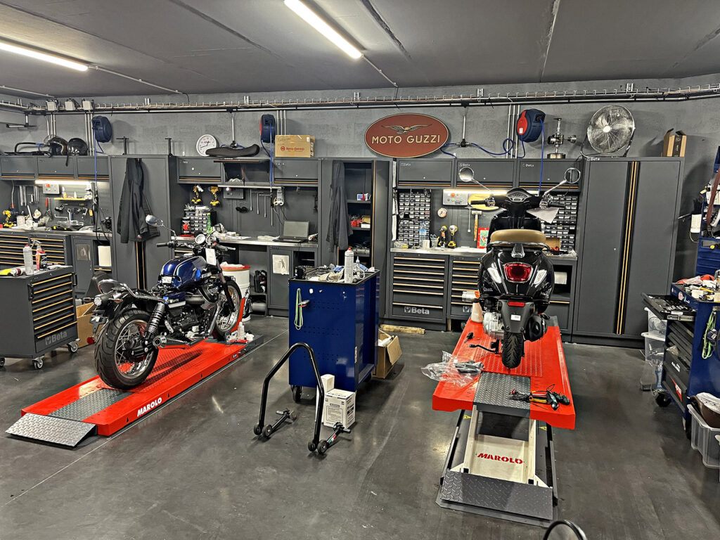 Un atelier de motocyclisme avec deux motos sur des ponts élévateurs rouges, entourées d'outils, d'armoires à outils et de divers équipements. Une enseigne "Moto Guzzi" est accrochée au mur du fond.