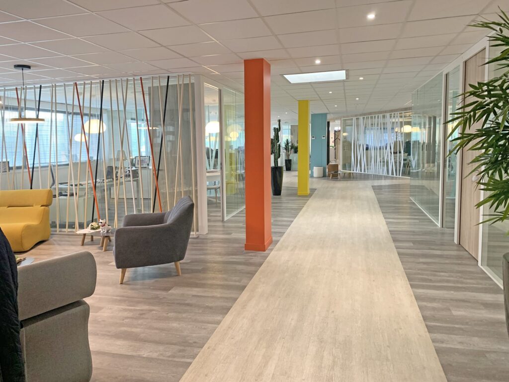 Intérieur de bureau moderne avec des salles de réunion vitrées, des piliers colorés, des sièges de salon et un long couloir avec un sol en bois.
