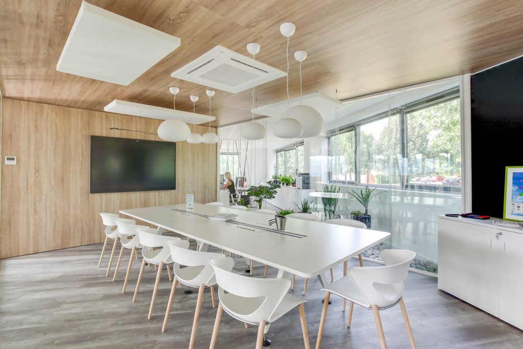 Salle de conférence moderne avec une longue table blanche, des chaises blanches, des lampes suspendues, un grand écran de télévision et des fenêtres allant du sol au plafond avec vue sur l'extérieur.