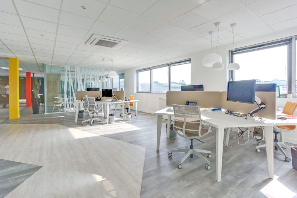 Bureau moderne à aire ouverte avec plusieurs bureaux, écrans d'ordinateur, chaises de bureau, grandes fenêtres et lumière vive. L'espace comporte des murs en verre et des accents colorés.