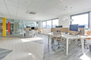 Bureau moderne à aire ouverte avec plusieurs bureaux, écrans d'ordinateur, chaises de bureau, grandes fenêtres et lumière vive. L'espace comporte des murs en verre et des accents colorés.