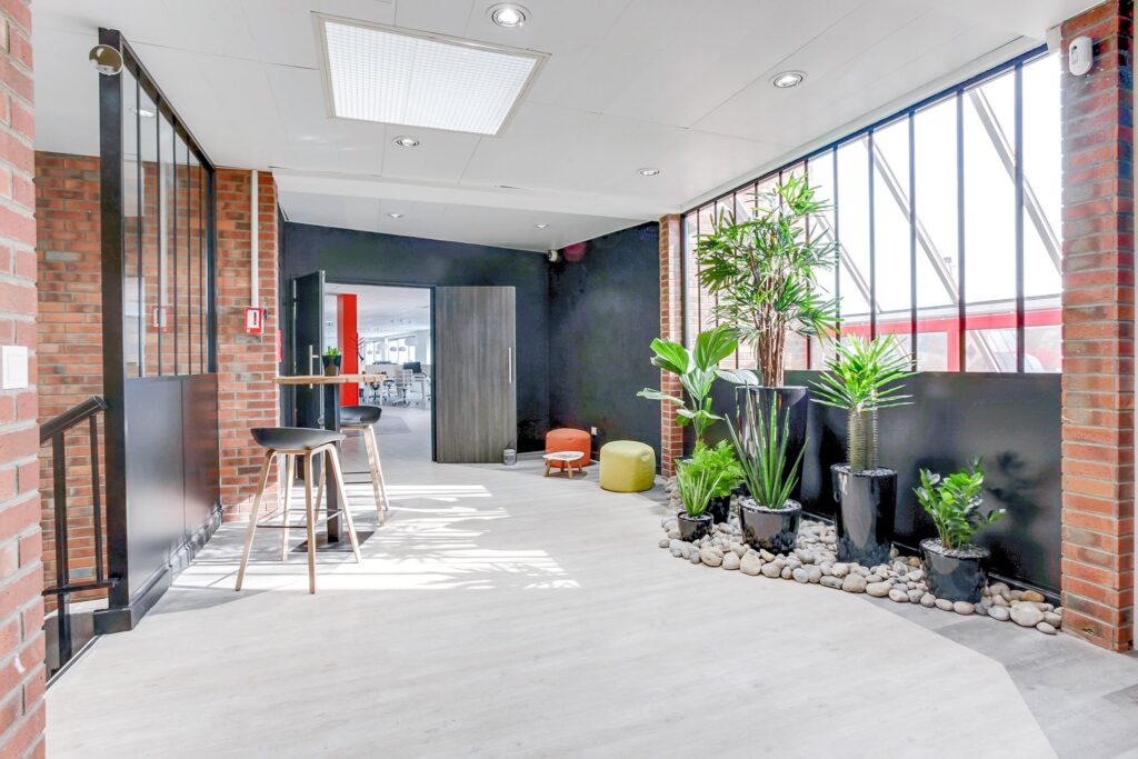 Salon de bureau lumineux avec des murs en briques, de hautes fenêtres, des tabourets de bar sur un comptoir, des plantes vertes en pot, un sol en bois clair et une vue sur un espace de travail adjacent.