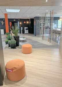 Salon de bureau moderne avec un sol en bois clair, des accents orange, des plantes dans des pots noirs, des poufs orange et un coin salon près de grandes fenêtres.