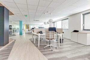 Espace de bureau moderne avec des bureaux blancs, des chaises, des ordinateurs et de grandes fenêtres qui laissent passer la lumière naturelle. La pièce est dotée d'un sol en bois clair et d'un design épuré et minimaliste.