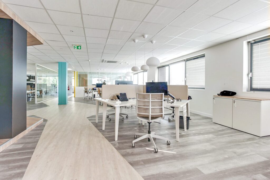 Bureau moderne à aire ouverte avec des bureaux blancs, des chaises ergonomiques, des ordinateurs et de grandes fenêtres laissant entrer la lumière naturelle ; l'espace semble propre et organisé.