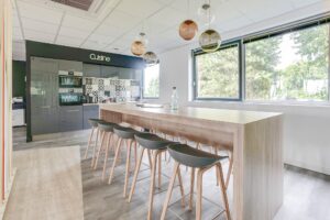 Cuisine moderne avec un long îlot en bois, six tabourets de bar, des lampes globe suspendues, des appareils encastrés et de grandes fenêtres laissant entrer la lumière naturelle.