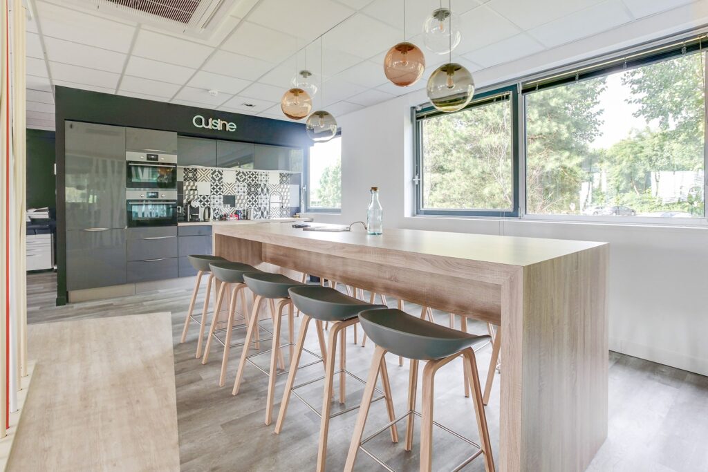 Cuisine moderne avec un grand îlot en bois, cinq tabourets de bar, des lampes suspendues, des appareils encastrés et une fenêtre donnant sur la verdure.