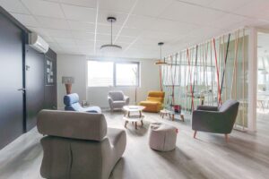 Salon de bureau moderne avec cinq fauteuils colorés, un ottoman rond, deux petites tables, de grandes fenêtres et une cloison en verre avec des tiges verticales colorées.