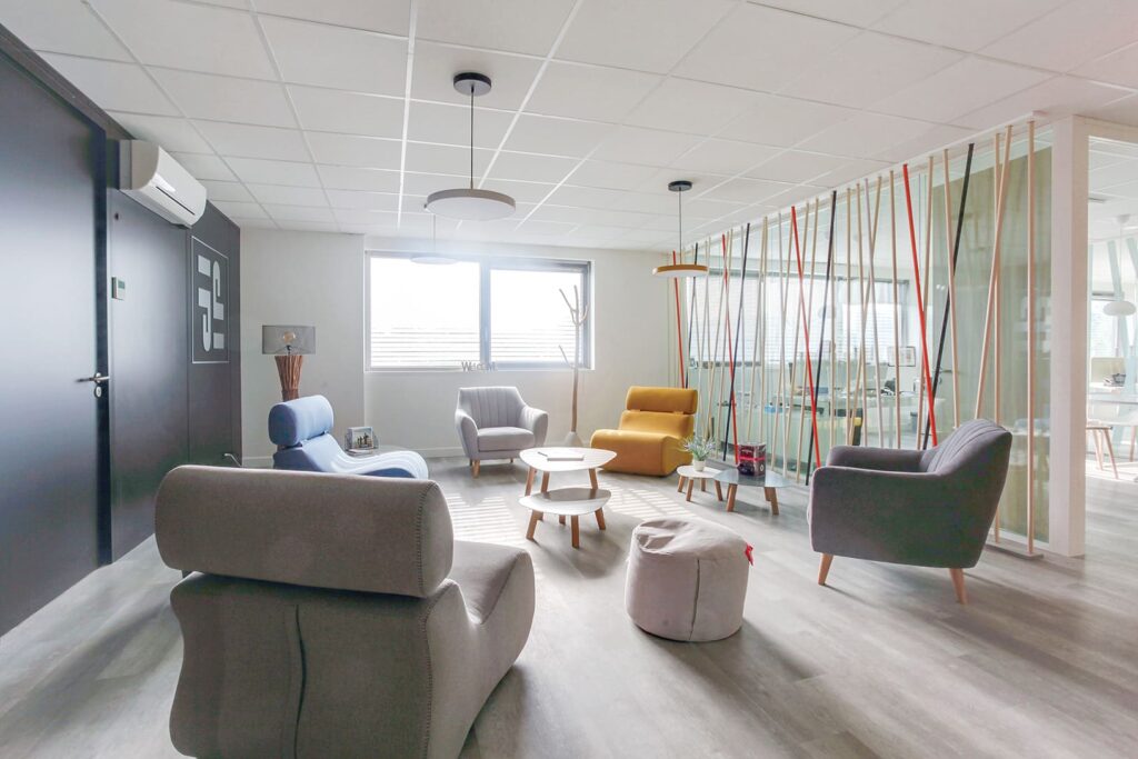 Salon de bureau moderne avec des fauteuils gris, jaunes et bleus, un pouf, de petites tables rondes, de grandes fenêtres et une cloison en verre avec des tiges verticales colorées.