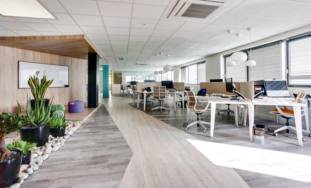 Bureau moderne à aire ouverte avec des bureaux, des chaises, des ordinateurs, des plantes en pot, de grandes fenêtres et un tableau blanc ; la lumière naturelle remplit l'espace.