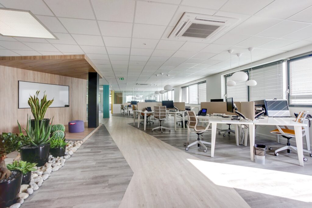 Bureau moderne à aire ouverte avec bureaux, chaises, ordinateurs, plantes d'intérieur, grandes fenêtres avec stores et tableau blanc sur le mur de gauche. L'espace est bien éclairé et bien rangé.