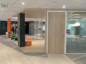 Intérieur de bureau moderne comprenant une salle de réunion en bois avec des murs en verre, des accents orange, des plantes d'intérieur et une table de conférence avec des chaises blanches et des lampes suspendues.