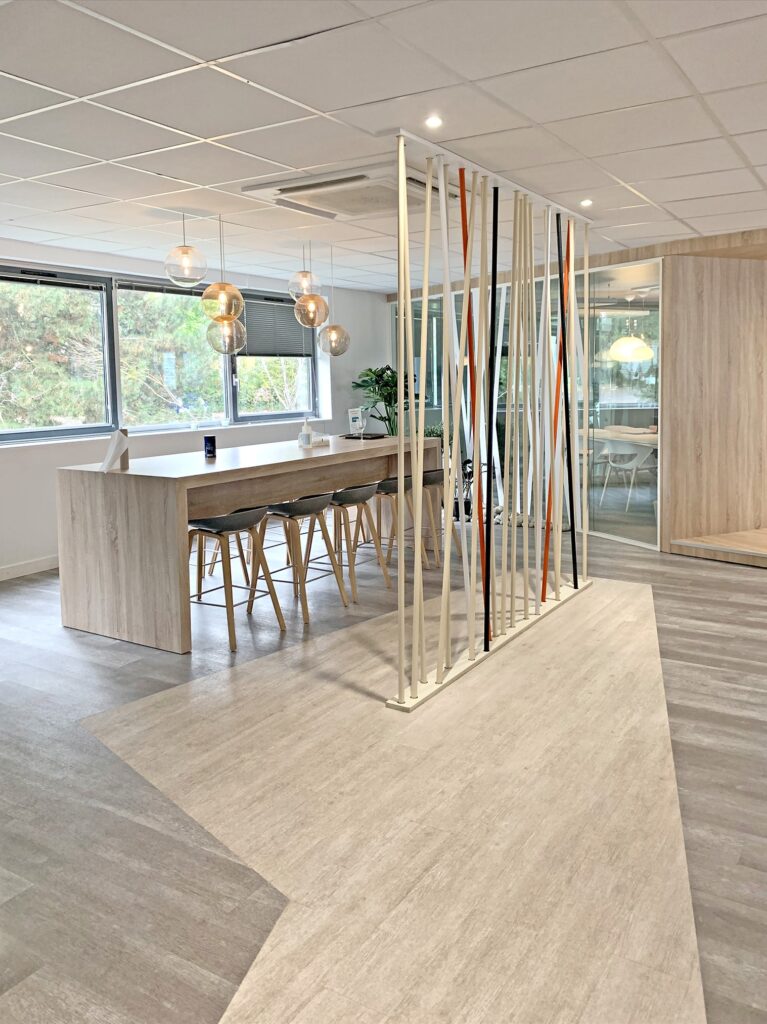 Bureau moderne avec une longue table en bois, des tabourets, des lampes suspendues, de grandes fenêtres et une cloison décorative avec des tiges verticales colorées.