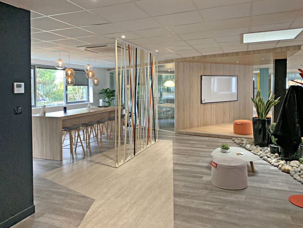 Bureau moderne avec une table commune, des tabourets de bar, des lampes suspendues, des plantes d'intérieur, un tableau blanc et des pierres décoratives au sol.