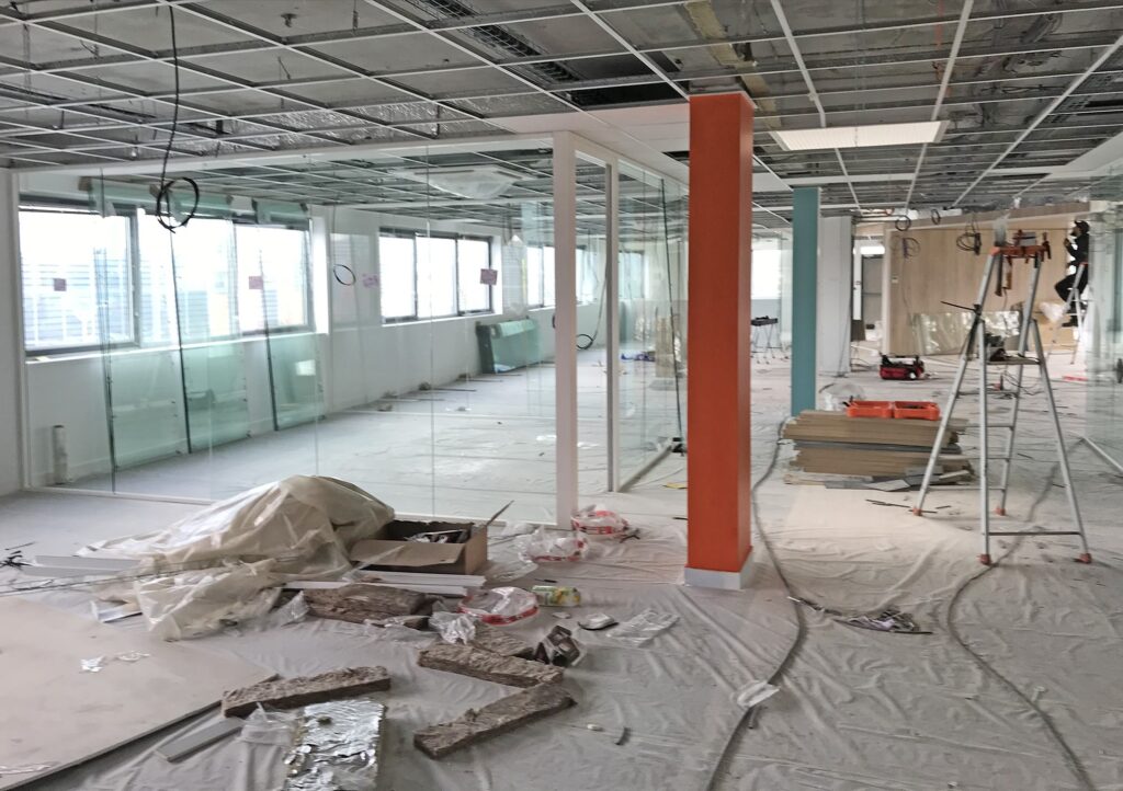 Un espace de bureaux en cours de rénovation avec des plafonds apparents, des matériaux de construction éparpillés, des cloisons en verre, des échelles et des revêtements de protection sur le sol.