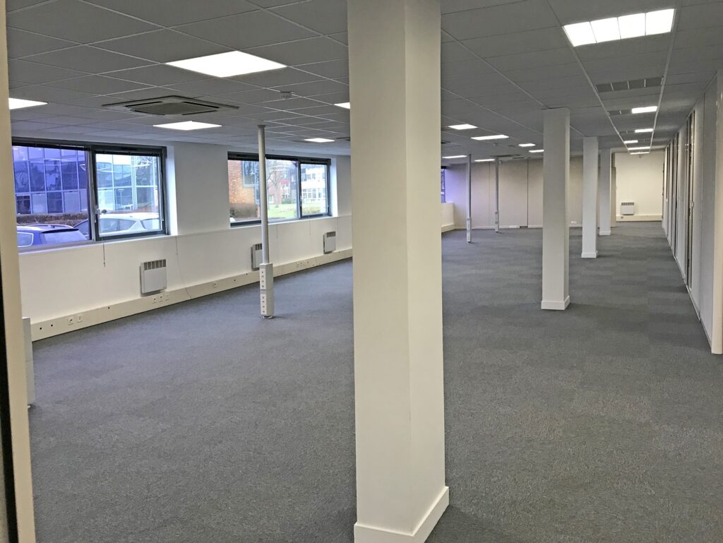 Un bureau vide avec une moquette grise, des murs blancs, de grandes fenêtres et plusieurs colonnes de soutien. Les plafonniers sont allumés et la pièce n'est pas meublée.