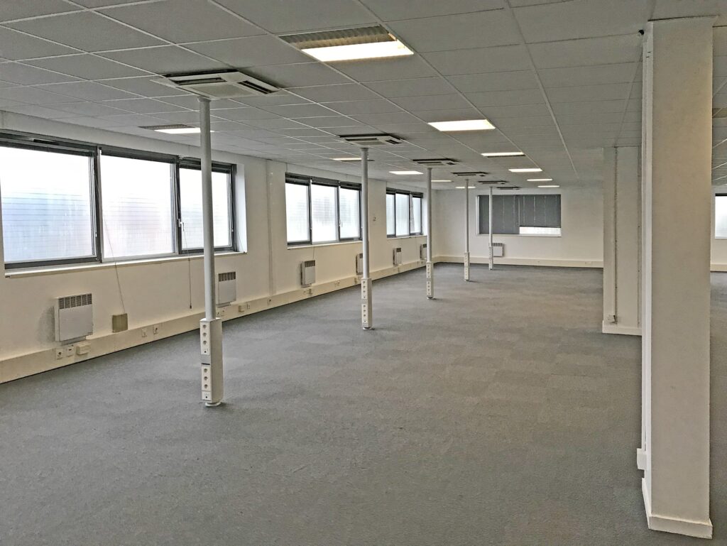 Un espace de bureau vide avec une moquette grise, des murs blancs, de grandes fenêtres et des plafonniers ; des colonnes de soutien avec des prises de courant courent au centre.