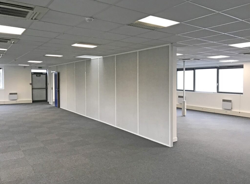 Un grand espace de bureau vide avec une moquette grise, des murs blancs, une cloison partielle et plusieurs fenêtres laissant entrer la lumière naturelle.