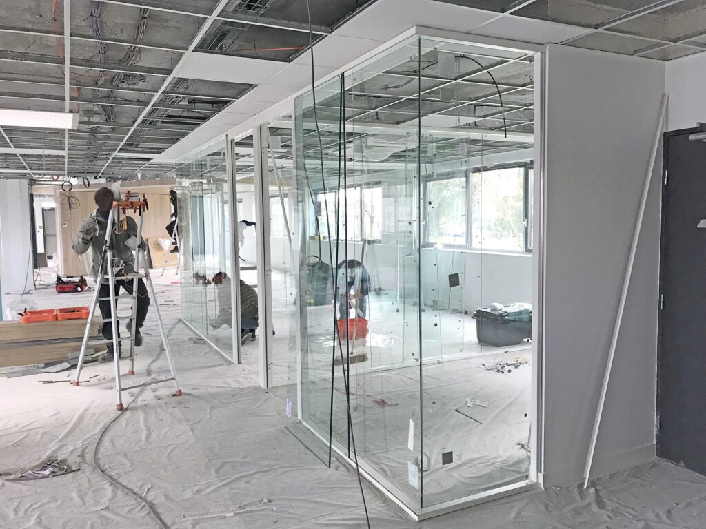 Ouvriers installant ou rénovant des cloisons de bureau en verre dans un bâtiment en construction, avec des outils et des matériaux éparpillés dans l'espace inachevé.