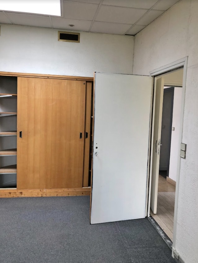 Une porte blanche bloque partiellement l'accès à un meuble coulissant en bois dans un bureau peu meublé et recouvert de moquette.