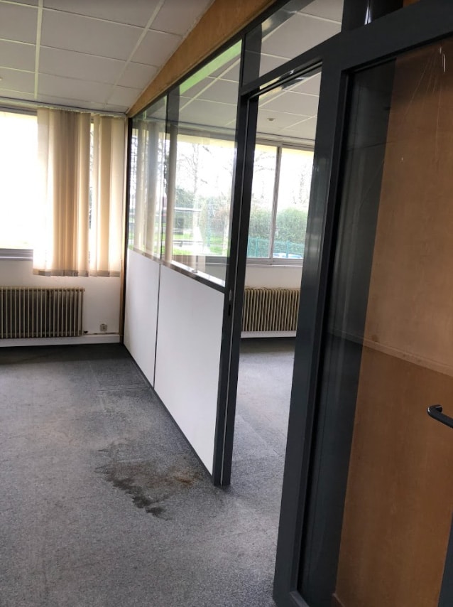 Un espace de bureau vide avec des cloisons en verre et en panneaux, un sol en moquette avec une tache visible, de grandes fenêtres et des stores verticaux.