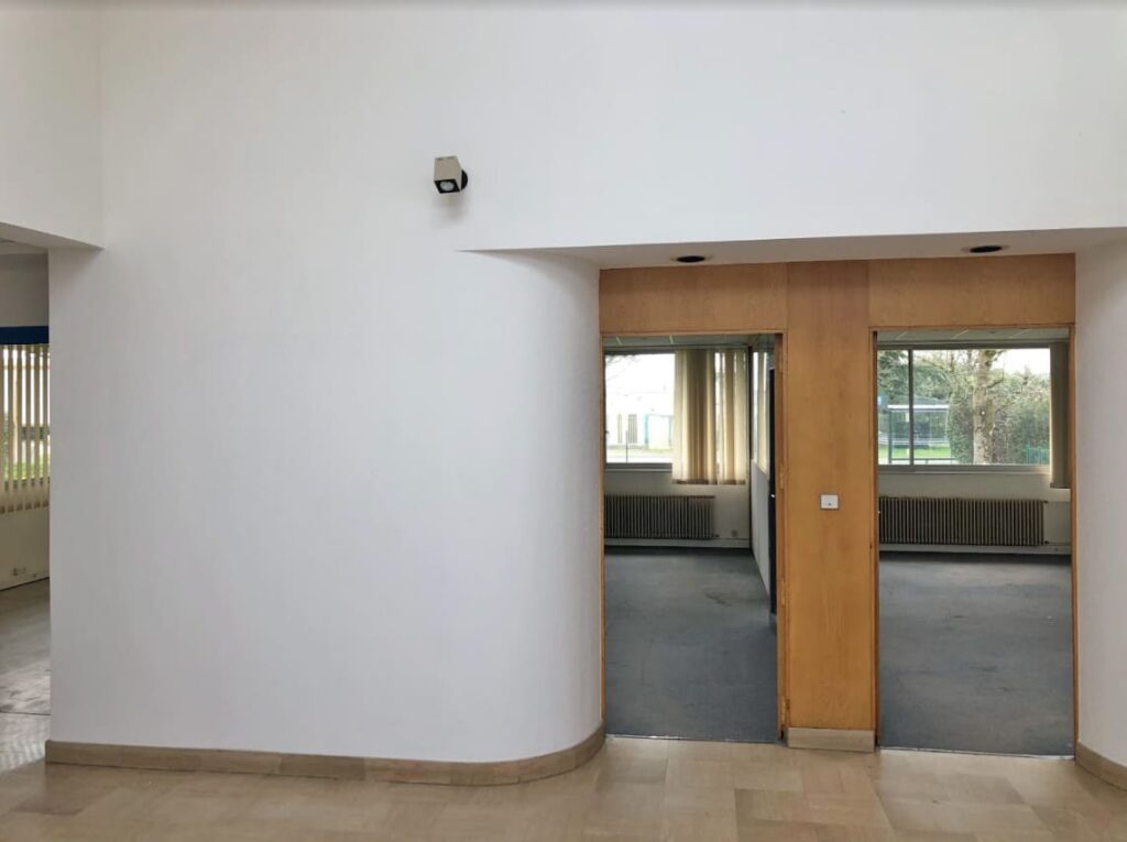 Une pièce aux murs blancs avec une double porte en bois mène à une autre pièce avec des sols recouverts de moquette et de grandes fenêtres montrant la verdure extérieure.