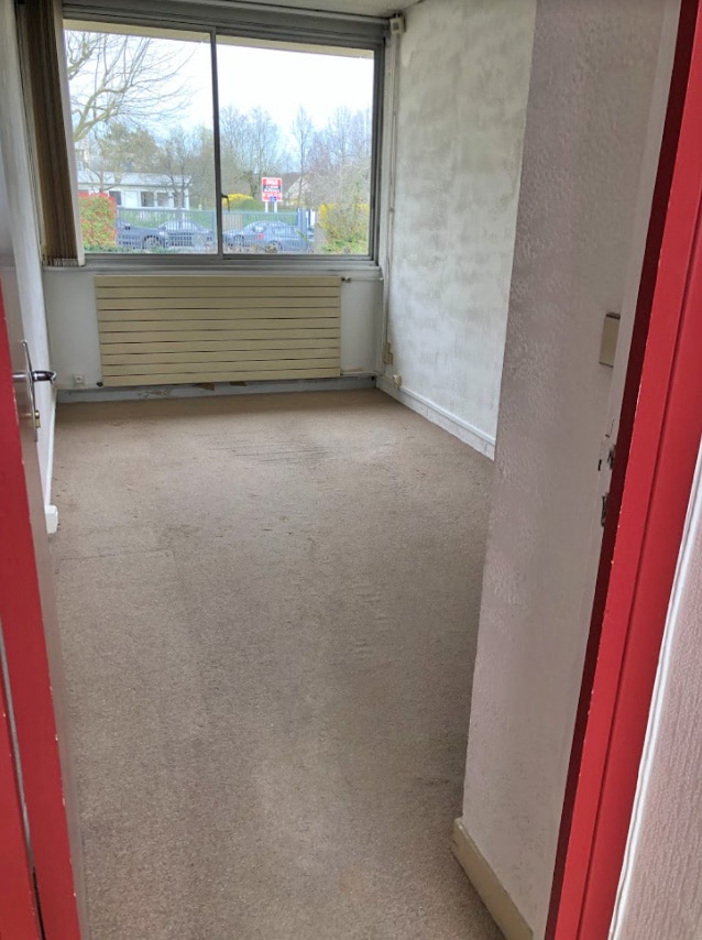 Petite pièce vide avec une moquette beige, des murs blancs et une grande fenêtre donnant sur un parking et des arbres, vue depuis une porte ouverte avec un cadre rouge.