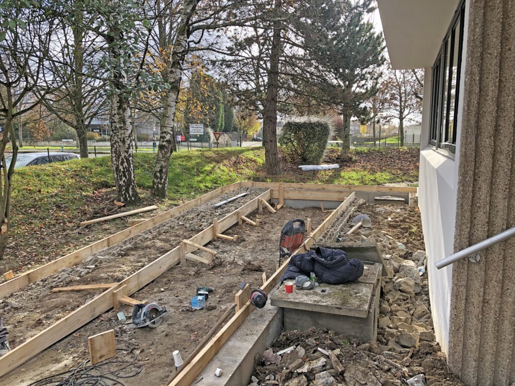 Chantier de construction avec une ossature en bois pour une fondation, des outils et des matériaux éparpillés, et des arbres en arrière-plan près de l'extérieur d'un bâtiment.