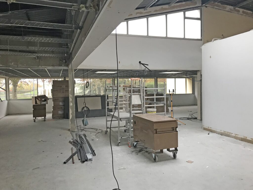 Grand espace intérieur en construction avec des échafaudages, des matériaux de construction et des débris éparpillés sur le sol. Le plafond exposé et les murs non finis sont visibles.